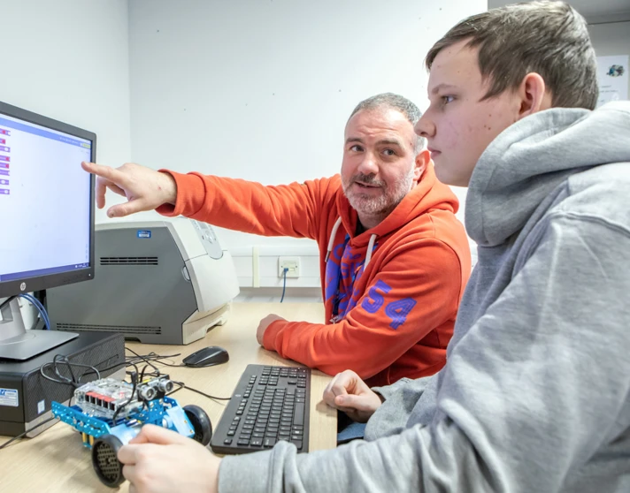 Lecturer in an orange hoodie guiding a student through programming a robotic vehicle on a computer screen in a computing classroom. Lecturer in an orange hoodie guiding a student through programming a robotic vehicle on a computer screen in a computing classroom.